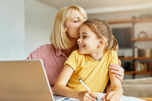 Woman whispers to daughter while hugging at a table with computer and homework
