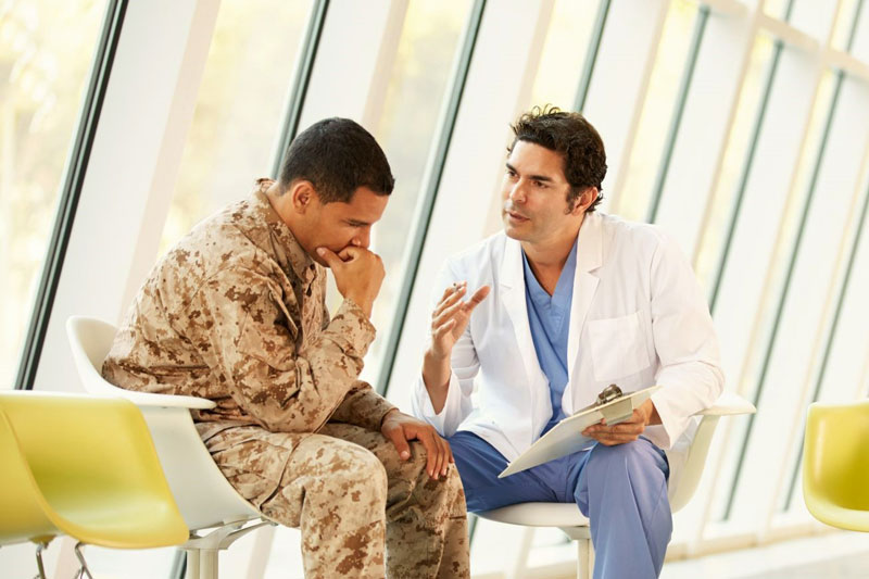 Doctor sits on bench with patient in military gear discussing treatment options