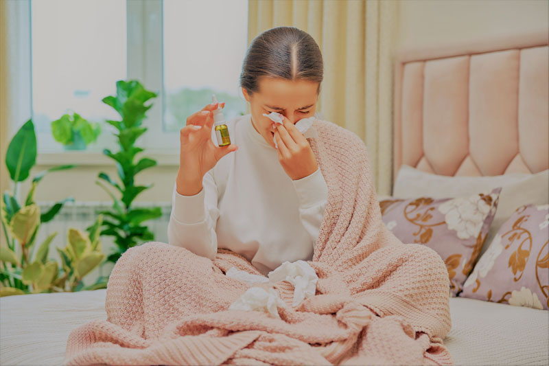 Woman sits on bed wrapped in blanket blowing her nose