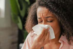 Indoors a woman holds a tissue to her nose as she winces with eyes closed