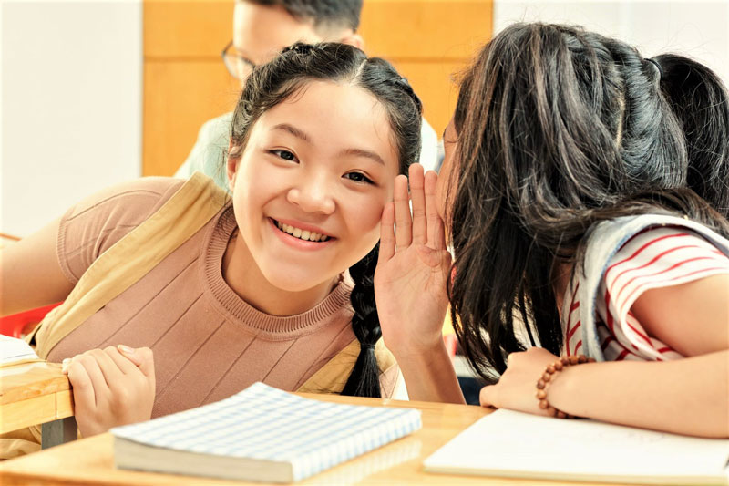 A female student whispers into the ear of another female student in their classroom