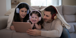 Parents and child with headphones on under a blanket, smiling, and watching a video on a laptop